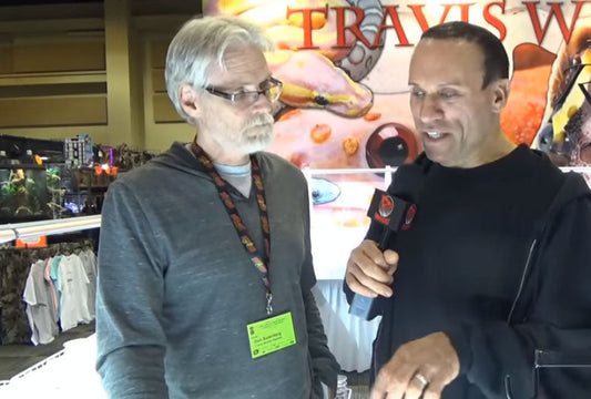 Two men in a reptile exhibit setting, one holding a microphone interviewing the other with reptile tanks and a colorful gecko poster in the background